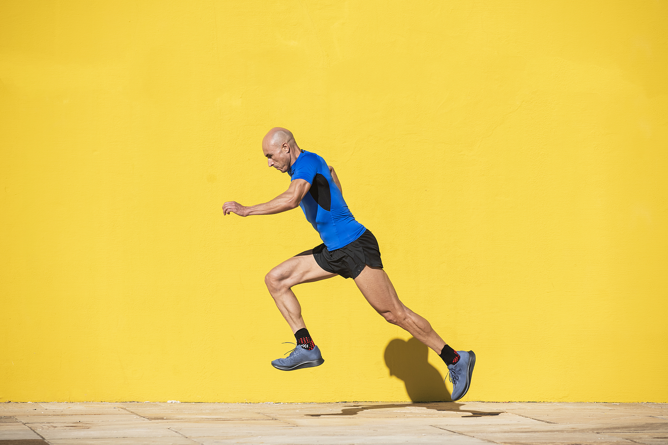 Sportsman with shaved head and sportswear starting a big step to run in the street in front of a yellow wall